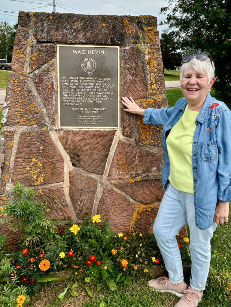 older white woman with white hair stands next to the Argyle Shore, PEI MacNevin monument, reading "We honour the memory of our MacNevin ancestors who emigrated from the Isles of Coll and Mull, Scotland, about 1810 AD to the shores of Prince Edward Island. They came from the known to the unknown with the dream of a better life for themselves and their descendants."